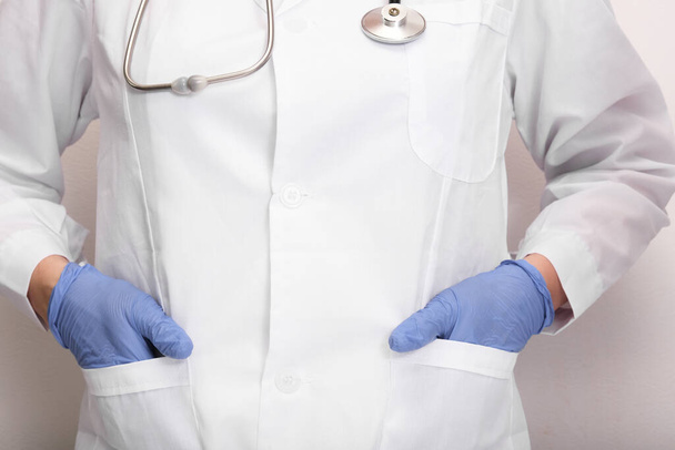 An unrecognizable doctor in white uniform and blue gloves with a stethoscope on neck holding hands in pockets close up - Foto, Imagem