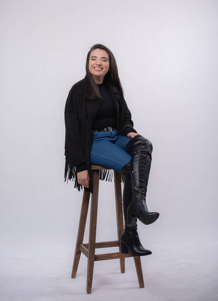 Happy beautiful girl model in black dress posing in studio isolated on white background,Teen girl in in blue jeans and black boots Full length portrait,sitting on a chair, - Foto, Imagen