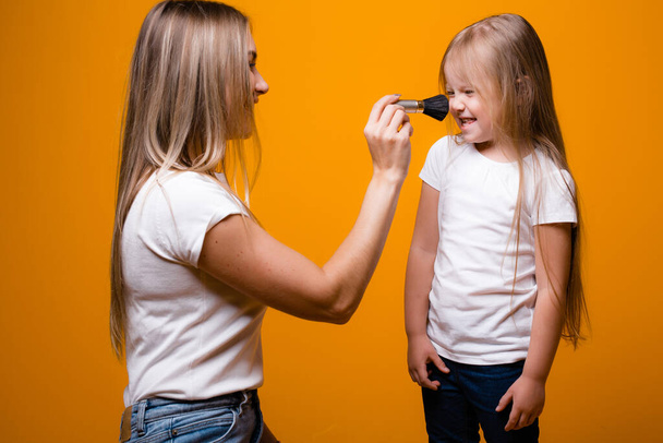 Mom teaches daughter to do makeup. Paints a girl blush with a brush on an orange background - Foto, Imagem