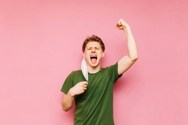 Happy young man with his mask removed rejoices at the end of the quarantine with a smile on his face, raises his hands in joy on a pink background.. Coronavirus pandemic. Quarantine. covid-19. - Foto, Bild