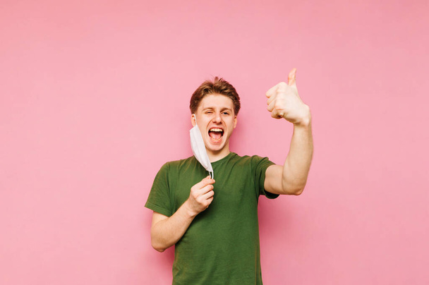 Ein positiver junger Mann steht mit abgenommener Schutzmaske auf rosa Hintergrund, blickt in die Kamera und lächelt, zeigt sein Gesicht und jubelt. Coronavirus-Pandemie. Quarantäne. covid-19. - Foto, Bild