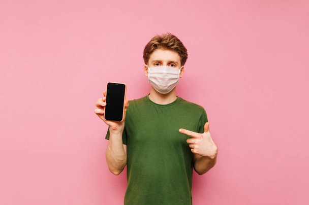 Handsome guy in a gauze mask stands with a smartphone in his hand on a pink background, points his finger at the black screen and looks at the camera with a serious face. - Photo, Image
