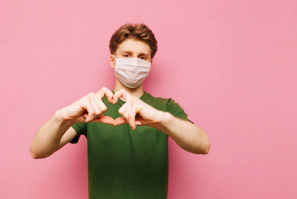 El tipo Cute está en un fondo rosa en una máscara médica y muestra un gesto cardíaco a la cámara. Isolado. Copie el espacio. pandemia de Coronavirus. COVID-19 - Foto, imagen