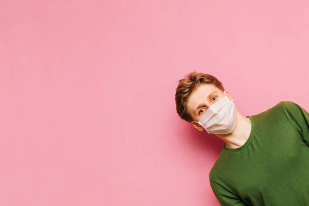 Смешной парень в медицинской маске на лице смотрит с правой стороны и смотрит в камеру, изолированную на розовом фоне. Коронавирус пандемический. COVID-19. Карантин. - Фото, изображение