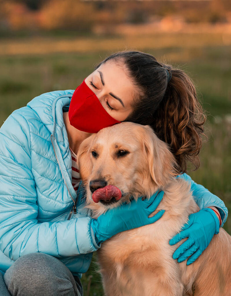 Žena v lékařsky chráněné masce obličeje se psem Zlatým retrívrem objímajícím. Pes hledící do kamery s lékařskou maskou. Domácí hygiena koncept - Fotografie, Obrázek