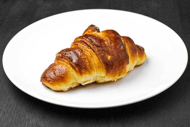  Čerstvý croissant s hnědou kůrkou na bílém talíři na černém stole. - Fotografie, Obrázek