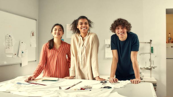 Tervezni jöttünk. Fiatal, vidám tervezők mosolyognak a kamerába, miközben együtt dolgoznak egy stúdióban. Kreatív Y-generációs szövetek, textilanyagok csoportja vágás előtt - Fotó, kép