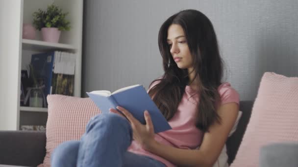 Beautiful attractive young woman reading a book while sitting on sofa at home - Filmati, video