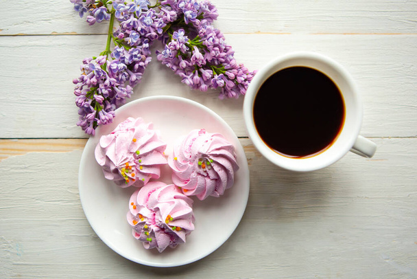Tasty meringue, coffee cup and lilac blooming branch on wooden table. Flat lay, top view - Photo, Image