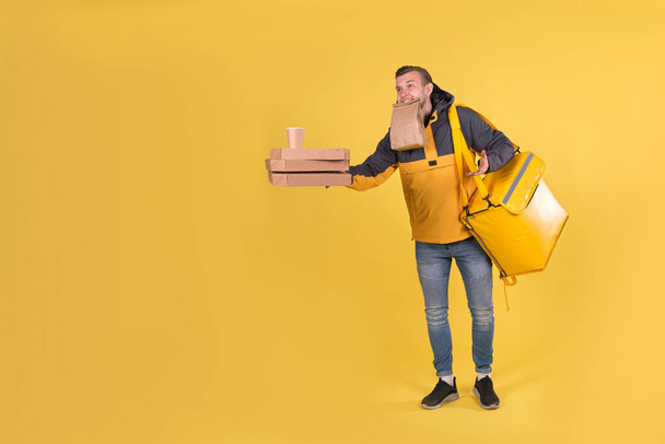 Portrait caucasian courier and food delivery young boy teenager in yellow uniform with thermo bag, offer forward an order with pizza and coffee cups in biodegradable eco-friendly packaging on yellow - Zdjęcie, obraz