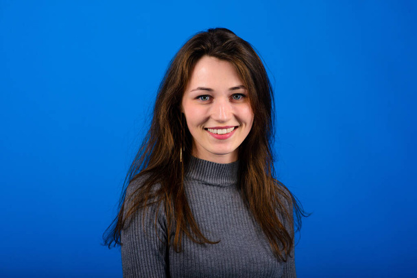 Foto einer lächelnden, fröhlichen jungen Frau in grauem Kleid auf blauem Hintergrund. Emotionales Porträt - Foto, Bild
