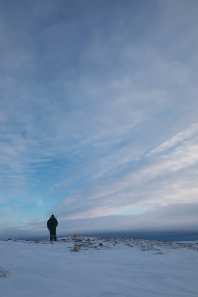 Man in winterkleding staand op sneeuw kijkend naar het weer op toendra landschap terwijl hij een geweer vasthoudt, nabij Arviat, Nunavut, Canada - Foto, afbeelding