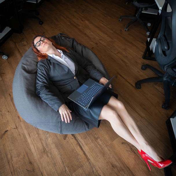 Modern advanced office. A woman in a business suit asleep in a beanbag while working on a laptop. Female manager is resting in a soft chair. Caring for the comfort of employees. - Foto, Imagem