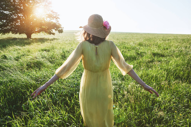 Pretty woman with yellow dress and hat walking in summer field in sunlght - Foto, afbeelding