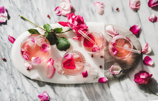 Θερινό δροσιστικό αναψυκτικό. Επίπεδη στρώση λεμονάδας τριαντάφυλλου με παγάκια σε ποτήρια και φρέσκα πέταλα τριαντάφυλλου σε οβάλ ταμπλό σερβιρίσματος πάνω από γκρι μαρμάρινο φόντο τραπεζιού, πάνω όψη - Φωτογραφία, εικόνα