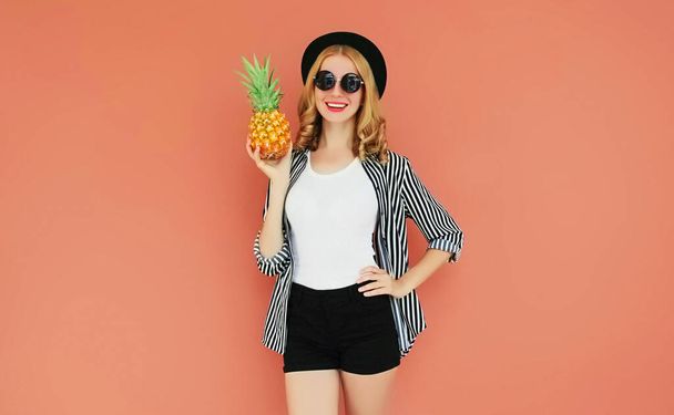 Portrait of happy young smiling woman with pineapple wearing a black hat, sunglasses, striped shirt over wall background - Photo, image