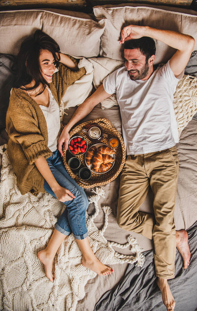 Young smiling happy couple lying on pastel colored blankets and having breakfast in bed with fresh croissants, strawberries and tea, top view. Comfort living, couples in love concept - Foto, Bild