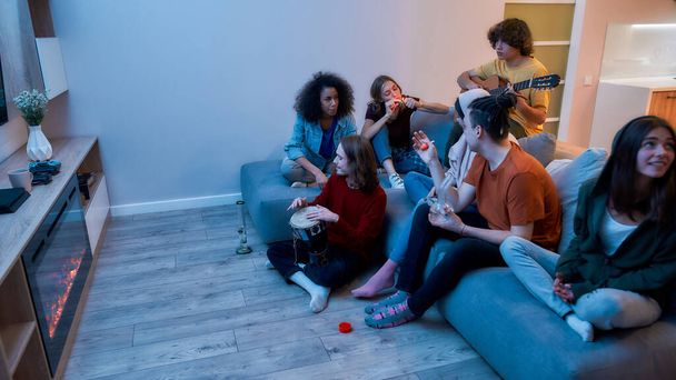 Young multicultural people smoking weed, marijuana together, playing music and relaxing on the sofa at home. Young guy lighting cannabis in the glass bong - Foto, imagen