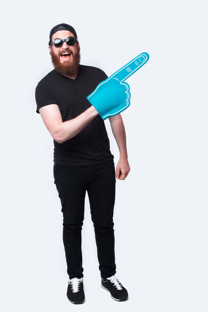 Full length photo bearded hipster man wearing sunglasses and pointing with big fan glove over white - Foto, immagini