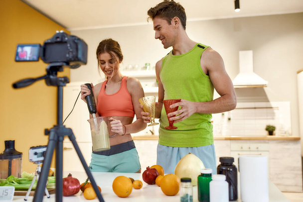 Experience. Create. Inspire. Young couple recording video blog or vlog about healthy nutrition on camera at home. Man and woman showing how to prepare smoothie, standing in the kitchen - Fotografie, Obrázek