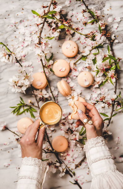 Flat-lay of womans hands holding sweet macaron cookies and fresh coffee in cup over white spring blossom flowers and white marble background, top view - Foto, afbeelding