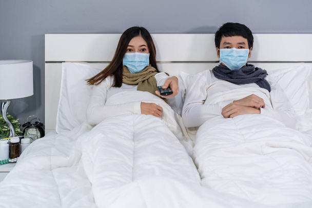 παντρεμένο ζευγάρι με ιατρικές μάσκες βλέποντας τηλεόραση σε ένα κρεβάτι, προστασία από τον κορωναϊό (covid-19) πανδημία έννοια. - Φωτογραφία, εικόνα
