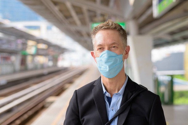 Portrait of mature businessman with mask for protection from corona virus outbreak at the sky train station - Foto, Imagen
