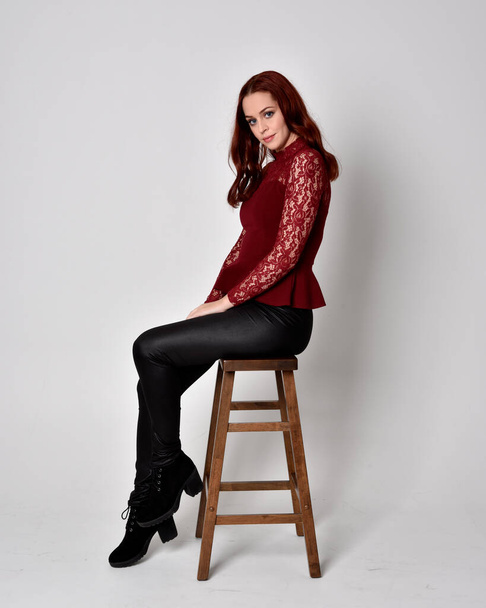 Portrait of a pretty girl with red hair wearing leather pants and long sleeved lace shirt.  full length sitting pose on wooden stool, isolated against a studio background - Zdjęcie, obraz