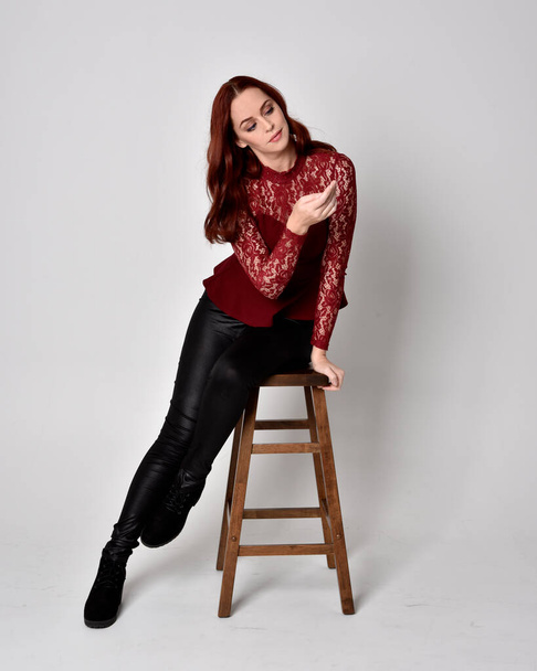 Portrait of a pretty girl with red hair wearing leather pants and long sleeved lace shirt.  full length sitting pose on wooden stool, isolated against a studio background - Foto, imagen