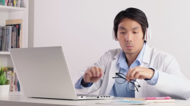 Aziatische man Doctor dragen hoofdtelefoon en witte jas of Gown en neem bril en het werken met laptop. Aziatische arts zie laptop monitor in ernstige emotie - Video
