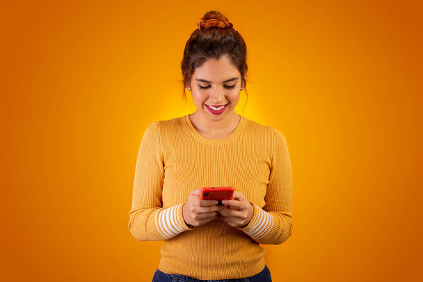Vrouw op gele achtergrond met behulp van mobiele telefoon en glimlachen - Foto, afbeelding