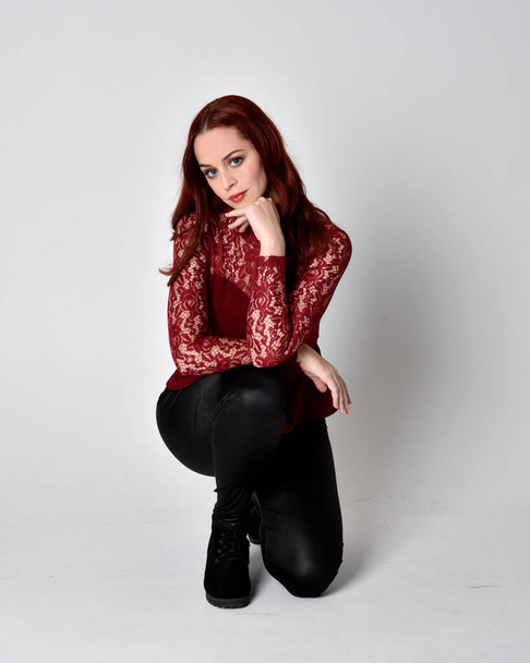 Portrait of a pretty girl with red hair wearing leather pants and long sleeved lace shirt.  full length kneeling pose  isolated against a studio background - Photo, image