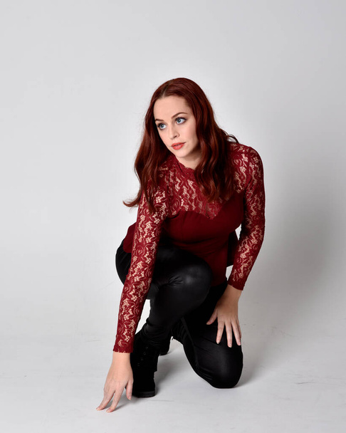 Portrait of a pretty girl with red hair wearing leather pants and long sleeved lace shirt.  full length kneeling pose  isolated against a studio background - Photo, Image