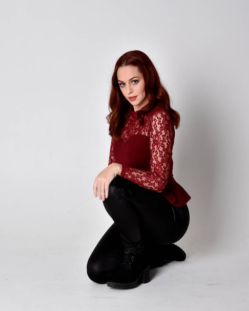 Portrait of a pretty girl with red hair wearing leather pants and long sleeved lace shirt.  full length kneeling pose  isolated against a studio background - Fotografie, Obrázek