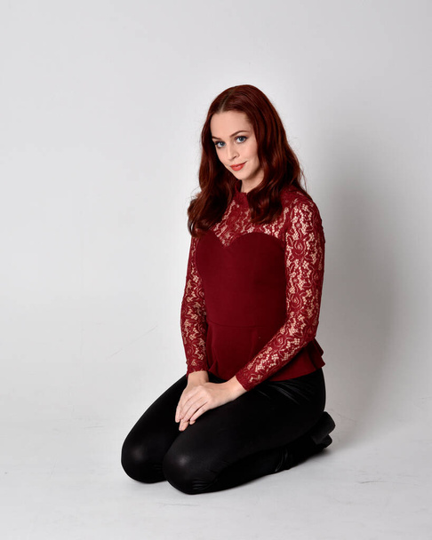 Portrait of a pretty girl with red hair wearing leather pants and long sleeved lace shirt.  full length kneeling pose  isolated against a studio background - Photo, Image