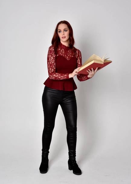 Portrait of a pretty girl with red hair wearing leather pants and long sleeved lace shirt.  full length standing pose holding a book,  isolated against a studio background. - Фото, зображення