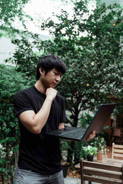 Egy fekete pólós férfi áll a kávézó kertjében a számítógépével a kezében.. - Fotó, kép