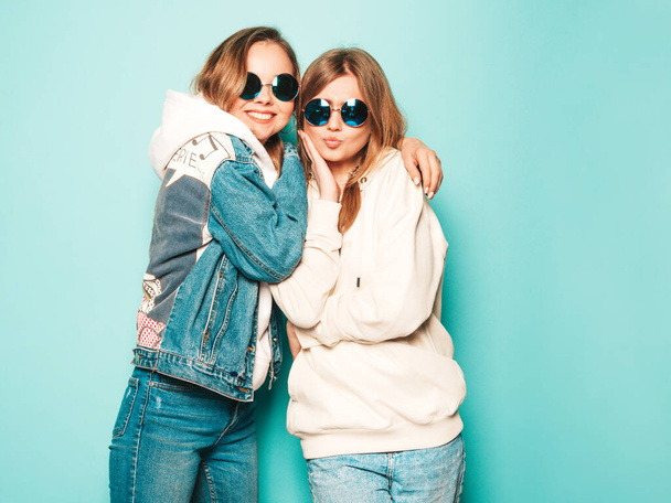Two young beautiful brunette smiling hipster girls in trendy summer hoodie and jeans jacket clothes. Sexy carefree women posing near blue wall. Trendy and positive models having fun in sunglasses - Photo, image