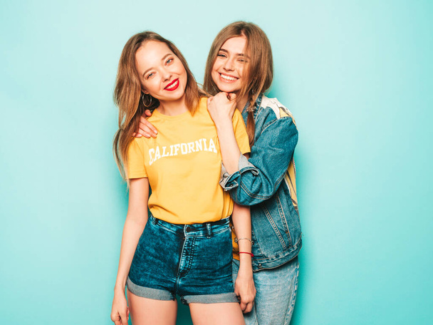 Two young beautiful smiling hipster girls in trendy summer yellow T-shirts and jeans jacket. Sexy carefree women posing near blue wall. Trendy and positive models having fun - Foto, Imagem