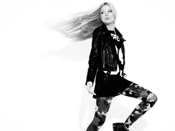 Young beautiful hipster girl in black leather jacket and stylish fashionable tight military pants leggings.Model with earring in nose.Sexy woman walking and jumping in studio.Flying hair in motion - Foto, Imagen