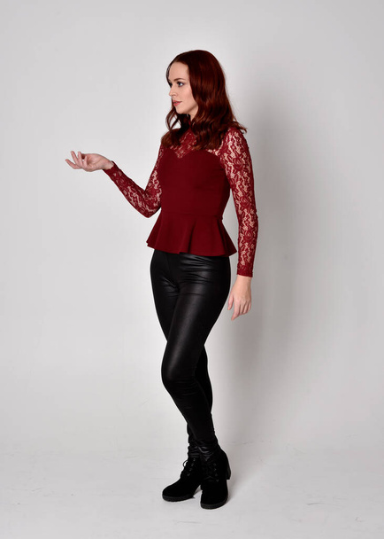 Portrait d'une jolie fille aux cheveux roux portant un pantalon en cuir et une chemise en dentelle à manches longues. pose debout pleine longueur avec geste de la main, isolé sur un fond de studio
. - Photo, image