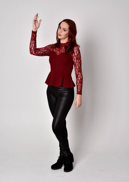 Портрет красивой девушки с рыжими волосами в кожаных штанах и кружевной рубашке с длинными рукавами. Стоячая поза с жестом руки, изолированная от студийного фона
. - Фото, изображение