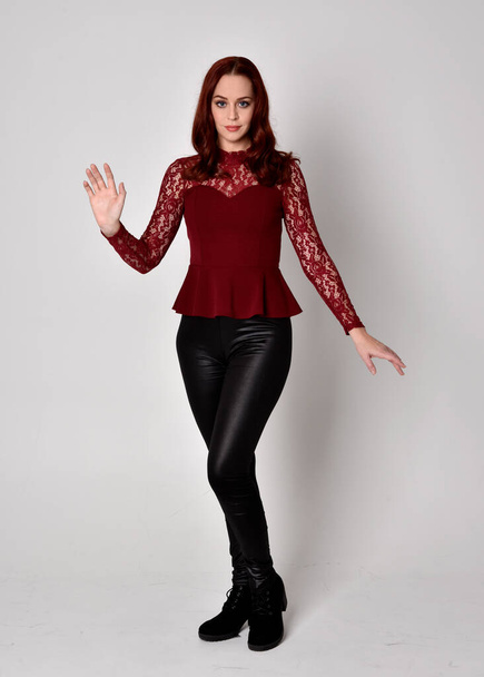 Portrait of a pretty girl with red hair wearing leather pants and long sleeved lace shirt.  full length standing pose with hand gesture, isolated against a studio background. - Photo, Image