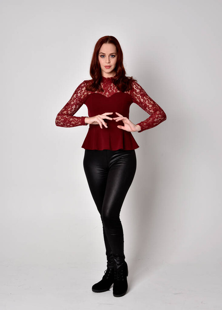 Portrait of a pretty girl with red hair wearing leather pants and long sleeved lace shirt.  full length standing pose with hand gesture, isolated against a studio background. - Foto, Imagem