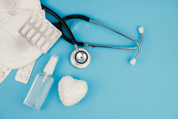 Fehér szív, orvosi sztetoszkóp, tabletták, maszkok és fertőtlenítő kék háttér.. - Fotó, kép