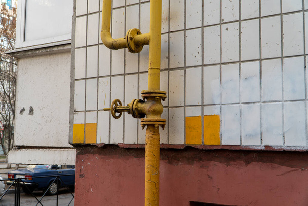Hauswand und verzweigte vertikale gelbe Gasleitung mit Ventil. Ventil mit Schwungrad am Schlauch erlaubt es, Kraftstoff zu verteilen und bei Bedarf auszuschalten. Wand mit weißen Fliesen. Horizontales Foto - Foto, Bild