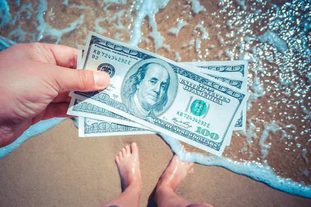 Девушка с деньгами в 300 долларов на фоне морских океанских волн с белой пеной и песчаным мокрым пляжем с ногами в песке крупным планом. Концепция финансирования отпуска деньги путешествия доллары отпуск - Фото, изображение