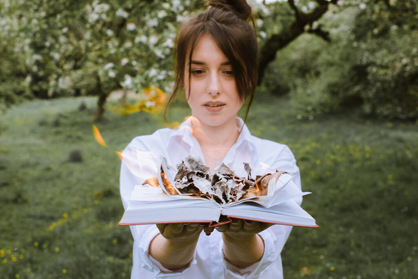 A lány világos ingben egy égő könyvet tart a kezében. Egy égő könyv a kezében. Egy könyv égett az erdőben. Egy fiatal nő az erdőben eléget egy könyvet. - Fotó, kép