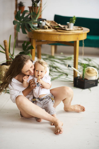 A young beautiful mom playing with her baby on the floor on near the wooden table with plants and green sofa - Fotografie, Obrázek