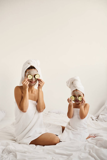 Μια νεαρή λευκή μητέρα και η μικρή της κόρη με τυλιγμένα μαλλιά σε λευκές πετσέτες μπάνιου διασκεδάζουν και απλώνουν κομμάτια αγγούρι στα μάτια τους..  - Φωτογραφία, εικόνα